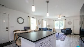 A modern kitchen with a central island, white cabinetry, and pendant lighting. There are two sofas and a television in the living area.