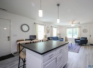 A modern kitchen with a central island, white cabinetry, and pendant lighting. There are two sofas and a television in the living area.