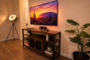 Una habitación con una mesa consola de madera, un televisor montado mostrando un atardecer desértico, una lámpara de pie y una planta en maceta.