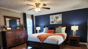 Een slaapkamer met een bed, een dressoir, nachtkastjes, een plafondventilator en een groot raam met jaloezieën.