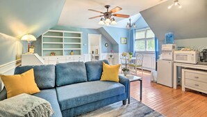 A modern living room with a blue sofa, yellow pillows, a ceiling fan, and a kitchen area with white appliances.