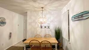 A dining area with a wooden table and chairs, a hanging light fixture, a clock, and a window with blinds. - Brigantine Condo w/ Gas Grill - Walk to Beach! (Brigantine)