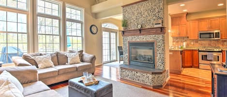 A spacious living room with a fireplace, a kitchen area with wooden cabinets, and large windows providing natural light.