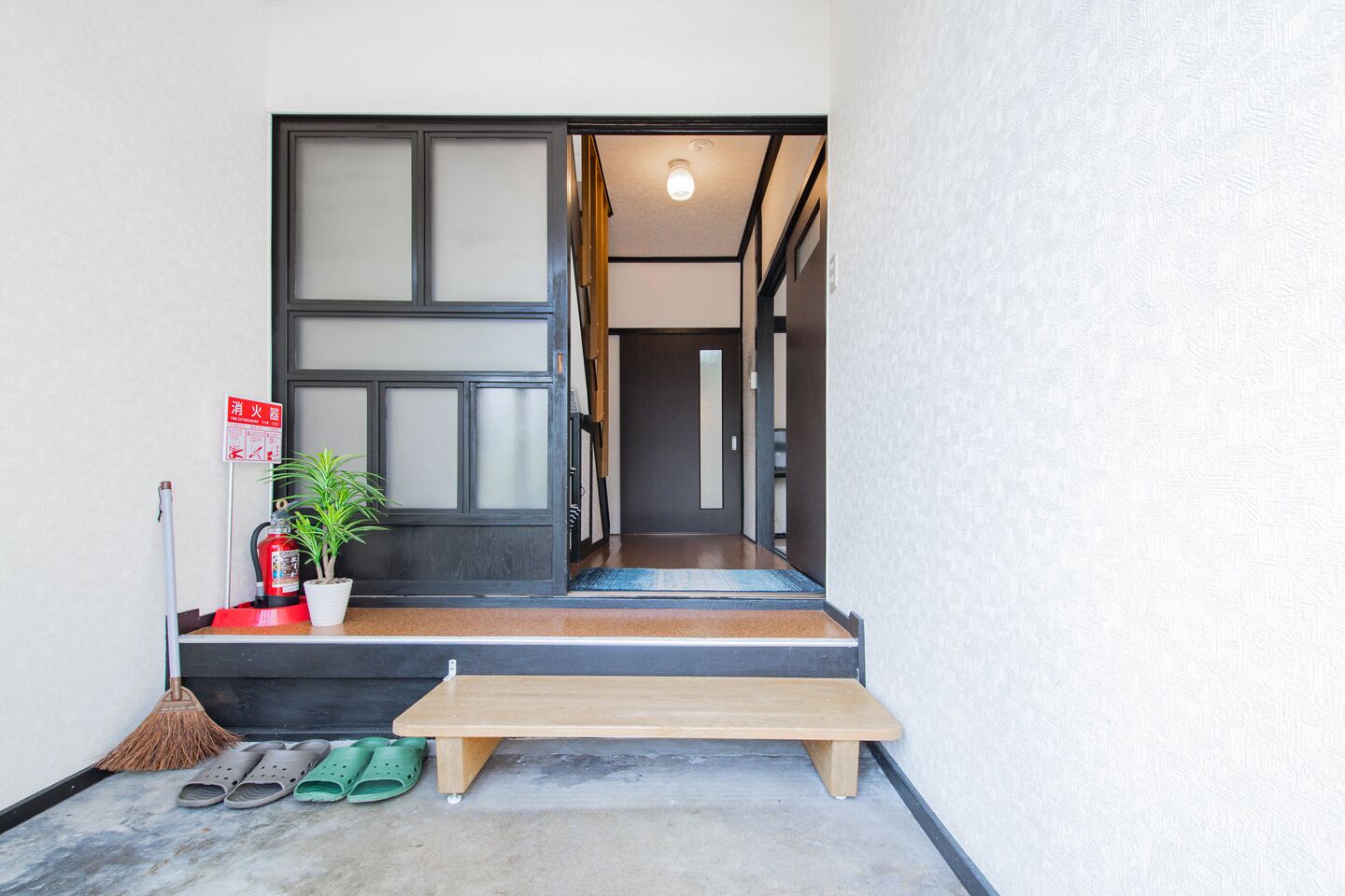 A wooden bench outside a building with a fire extinguisher, a broom, and a potted plant on the steps.