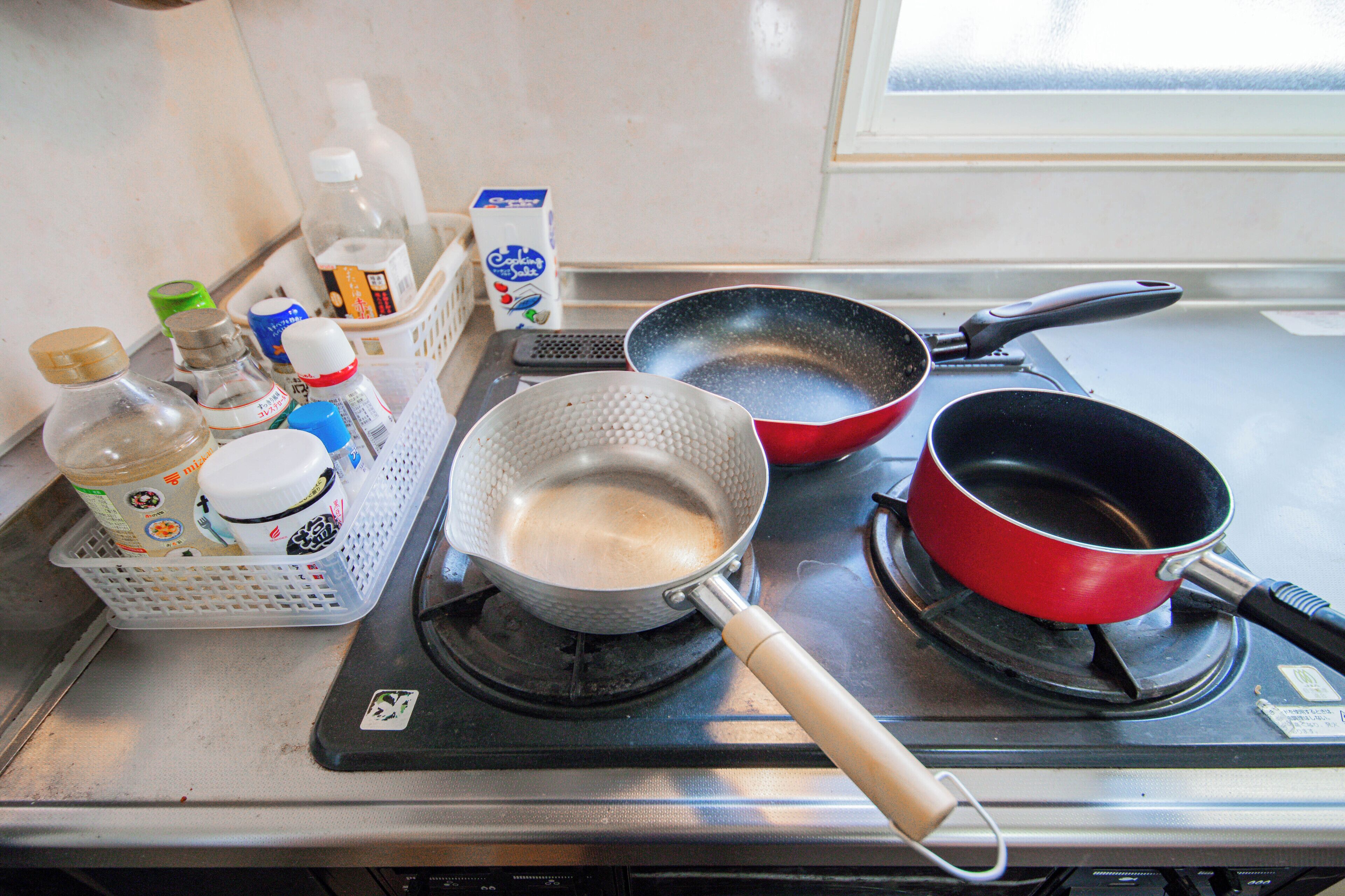 Una cocina con estufa, dos sartenes, un rallador y diversos utensilios de cocina sobre la encimera.