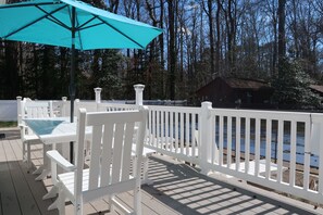 Eine Holzterrasse mit weißen Stühlen und einem türkisblauen Sonnenschirm, mit Blick auf ein Gewässer und ein bewaldetes Gebiet.