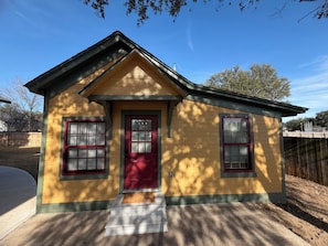 A small yellow house with a red door and windows, a front porch with a welcome mat, and a wooden fence in the background.