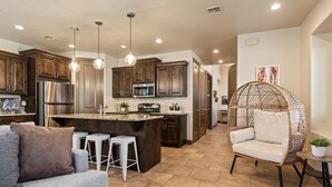 A modern kitchen with wooden cabinets, stainless steel appliances, and a central island. There is a seating area with a sofa and a wicker chair.