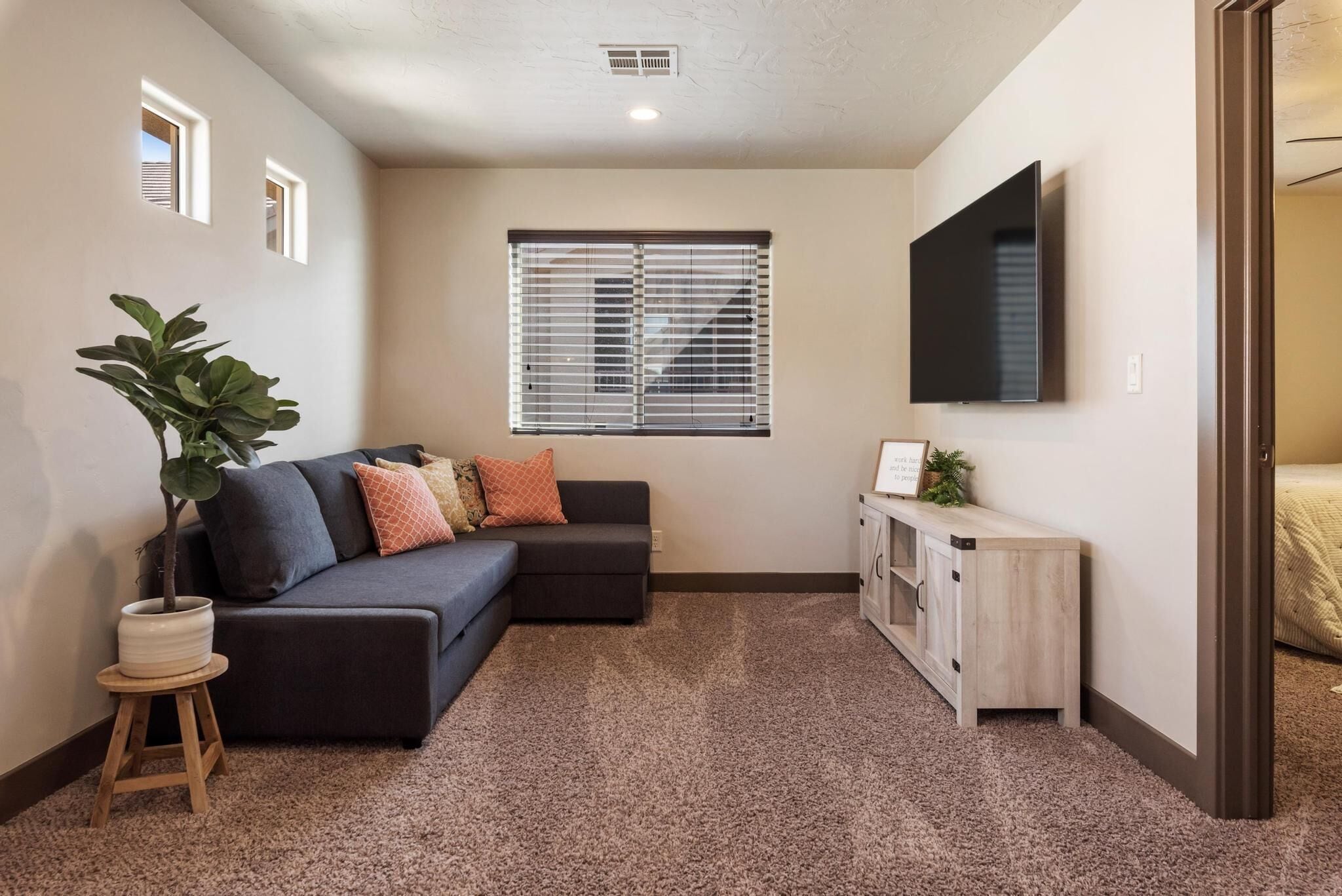 A cozy living room with a dark sofa, a small side table, a potted plant, a window with blinds, a TV mounted on the wall, and a wooden cabinet.