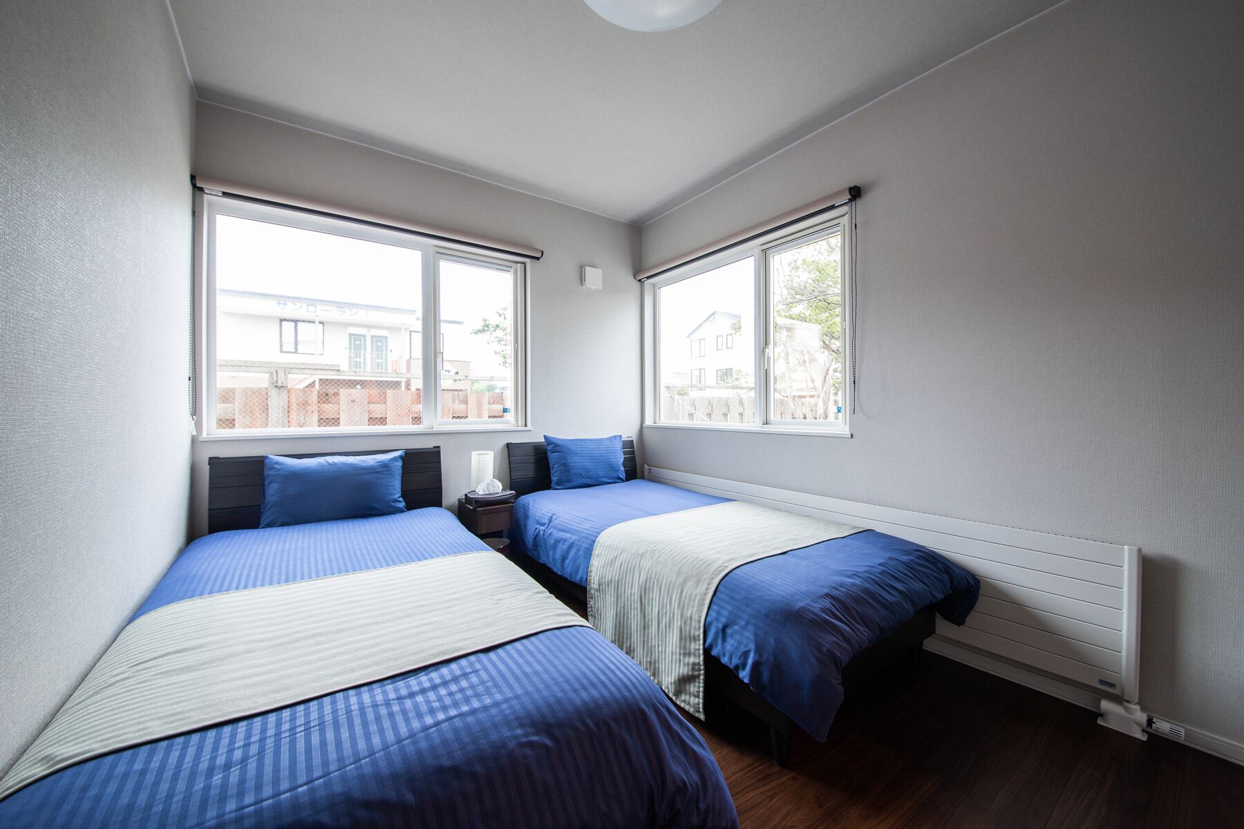 Un dormitorio con dos camas, cada una con ropa de cama azul y una manta blanca, una mesita de noche con una lámpara y una ventana con vistas a una casa.