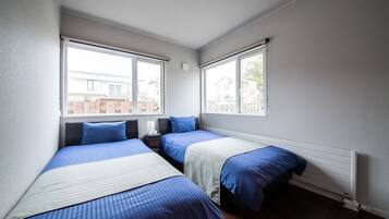 Un dormitorio con dos camas, cada una con ropa de cama azul y una manta blanca, una mesita de noche con una lámpara y una ventana con vistas a una casa.