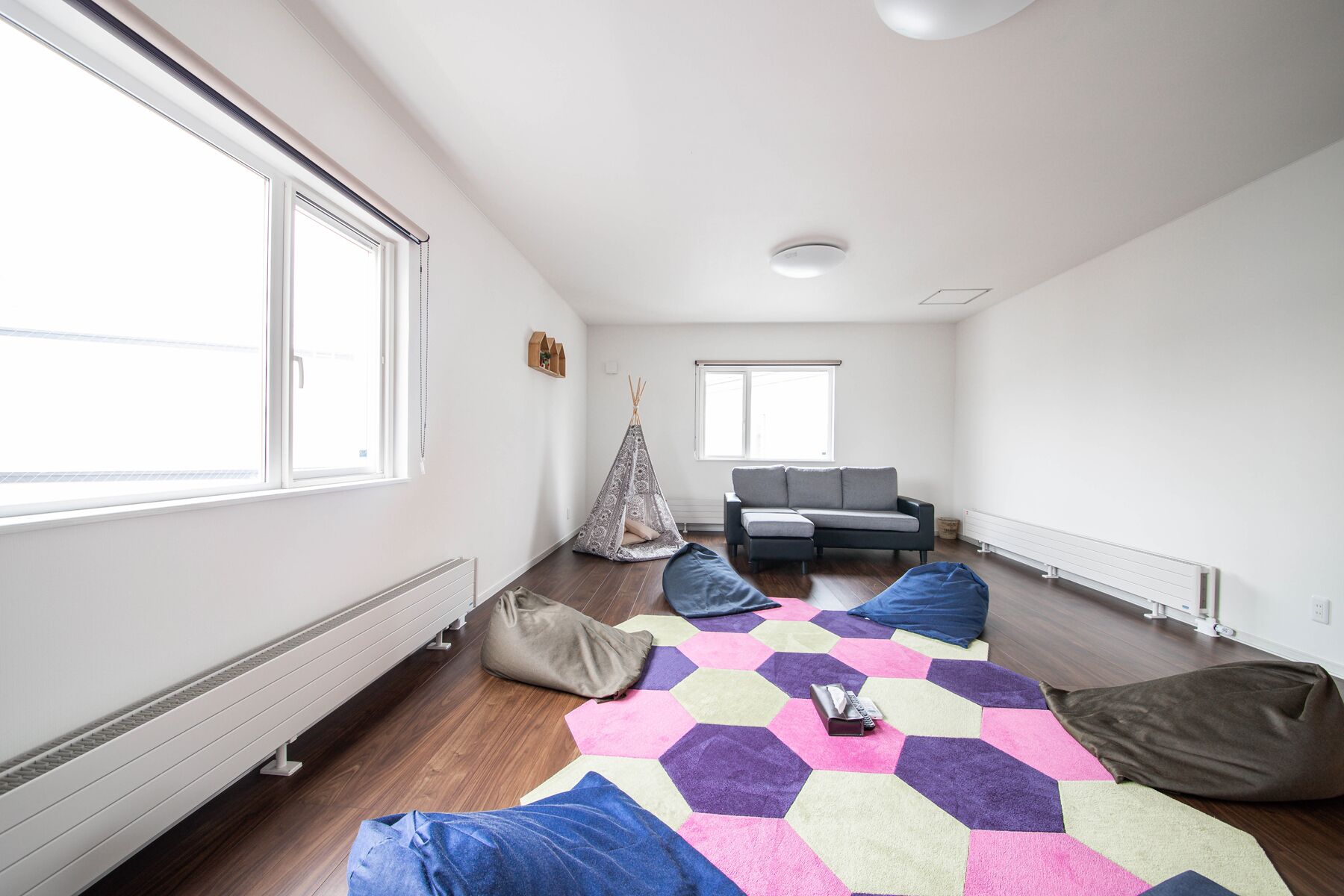 A modern, well-lit room with a large window, a sofa, a small tent, and a colorful hexagonal rug.