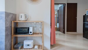 A compact kitchen with a microwave, toaster, and kettle on a wooden shelf, a wall clock, and a door leading to another room. - CLASSE TROIS (Sapporo)