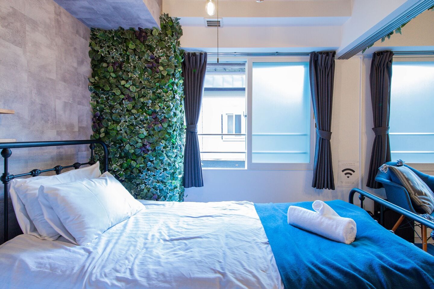 A hotel room with a bed, a blue blanket, a white pillow, a towel, a window with curtains, and a wall covered with green foliage.