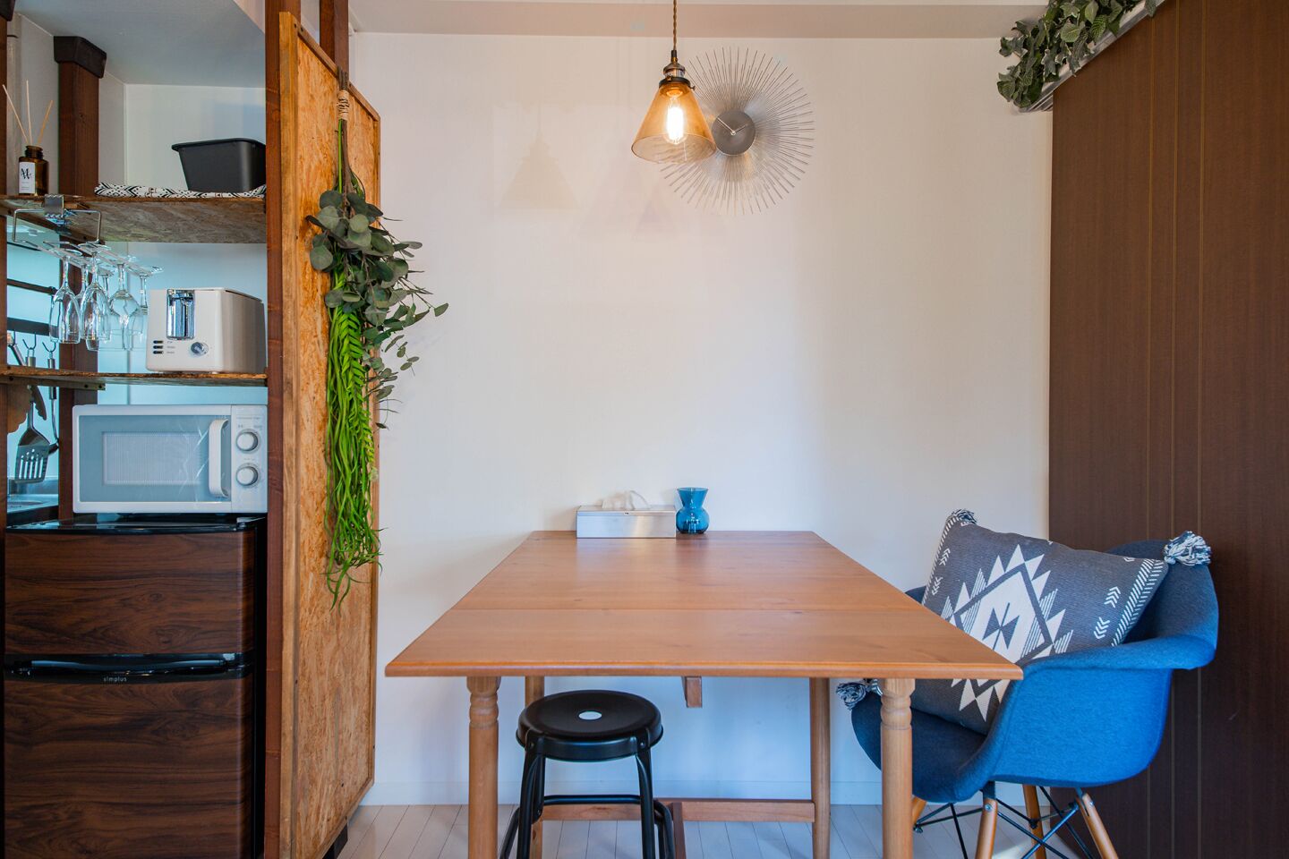A compact kitchen with a wooden table, a blue chair, and a hanging light fixture.
