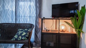 Ein Zimmer mit Fernseher auf einem Schrank, ein Korbstuhl mit Kissen, einer Pflanze und einem Schrank mit Lampe.