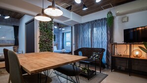 Ein modernes Wohnzimmer mit einem Holzesstisch, schwarzen Stühlen, einem schwarzen Sofa, einem großen Fernseher und einer dekorativen Pflanzenwand.