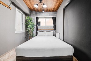 Una habitación pequeña con cama, ventana con cortinas, radiador y una planta.