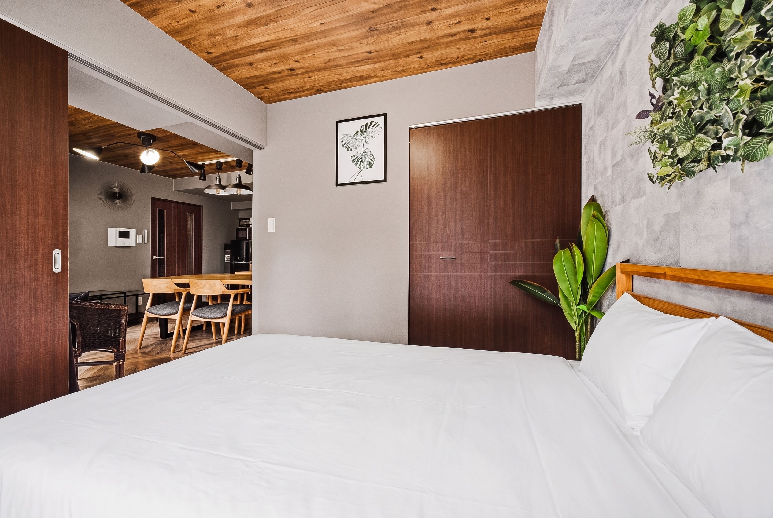A bedroom with a wooden ceiling, a bed with white bedding, a dining area with a wooden table and chairs, and a wall-mounted plant.