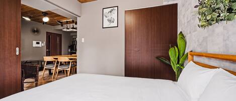 A bedroom with a wooden ceiling, a bed with white bedding, a dining area with a wooden table and chairs, and a wall-mounted plant.