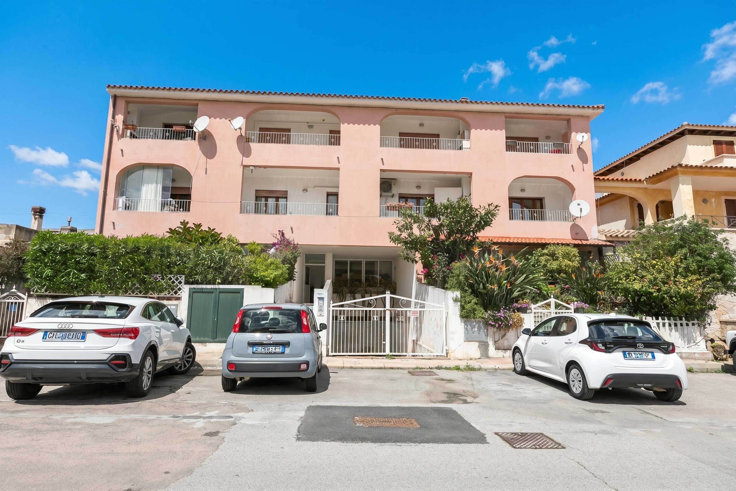 Un edifici de diverses plantes amb balcons, una entrada tancada amb reixa i cotxes aparcats.