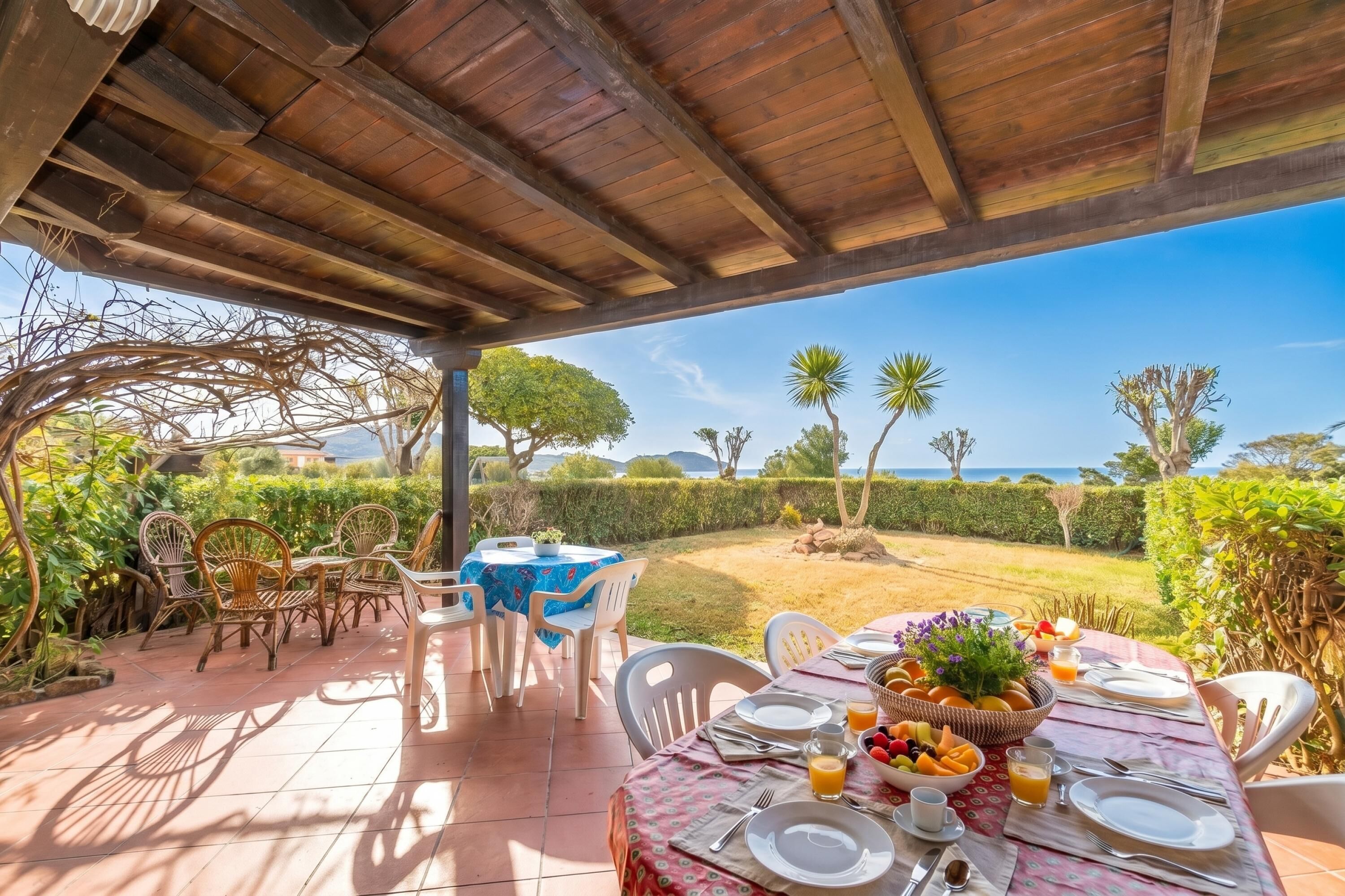 A covered outdoor dining area with tables and chairs, a view of the sea, and a garden with various plants.