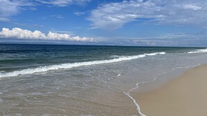 Une plage où les vagues viennent lécher le rivage, un ciel dégagé et des montagnes à l’horizon.