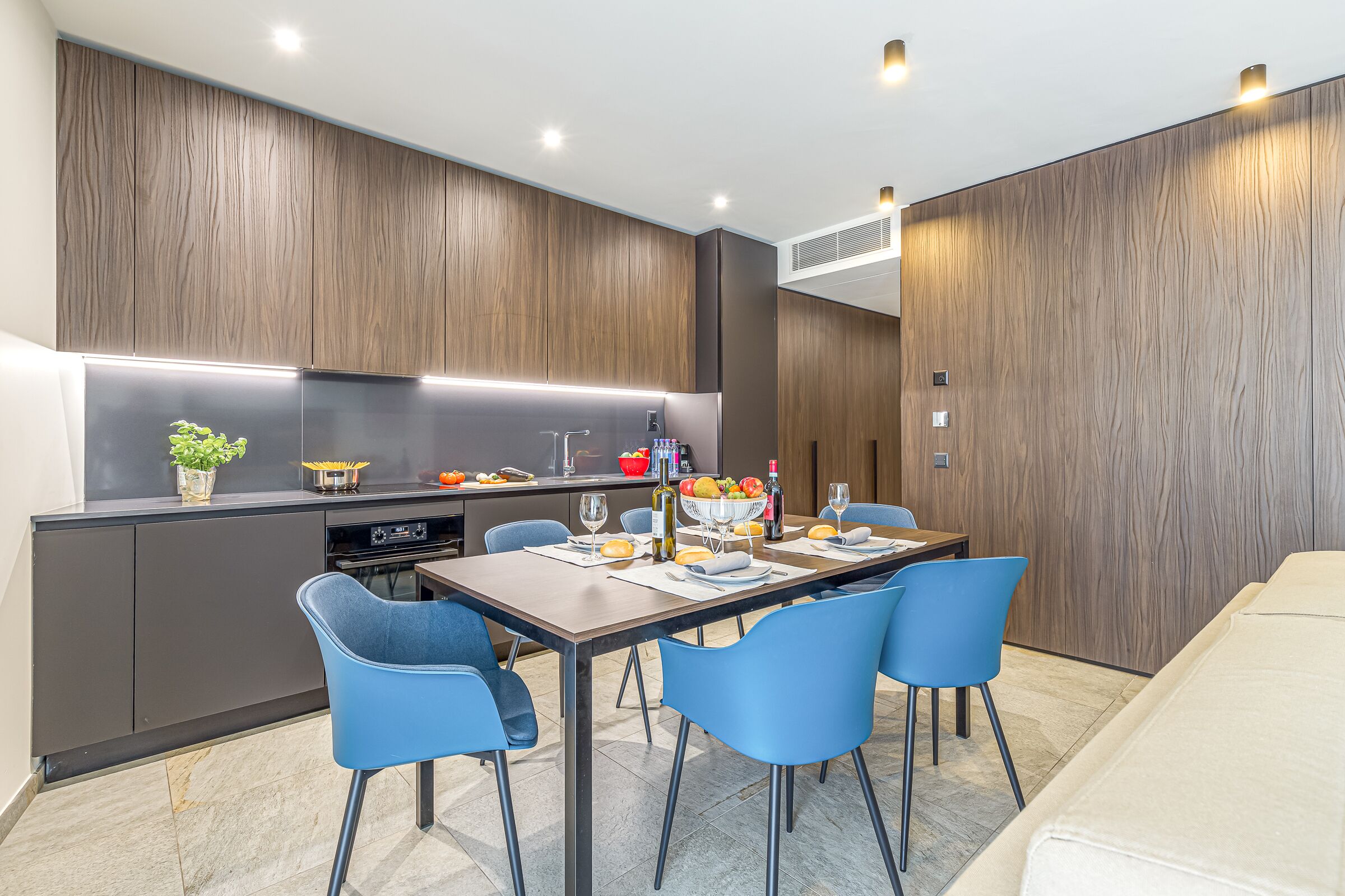 Una cucina moderna con mobili in legno scuro, un'isola centrale e un'area da pranzo con sedie blu e un tavolo imbandito per quattro persone.