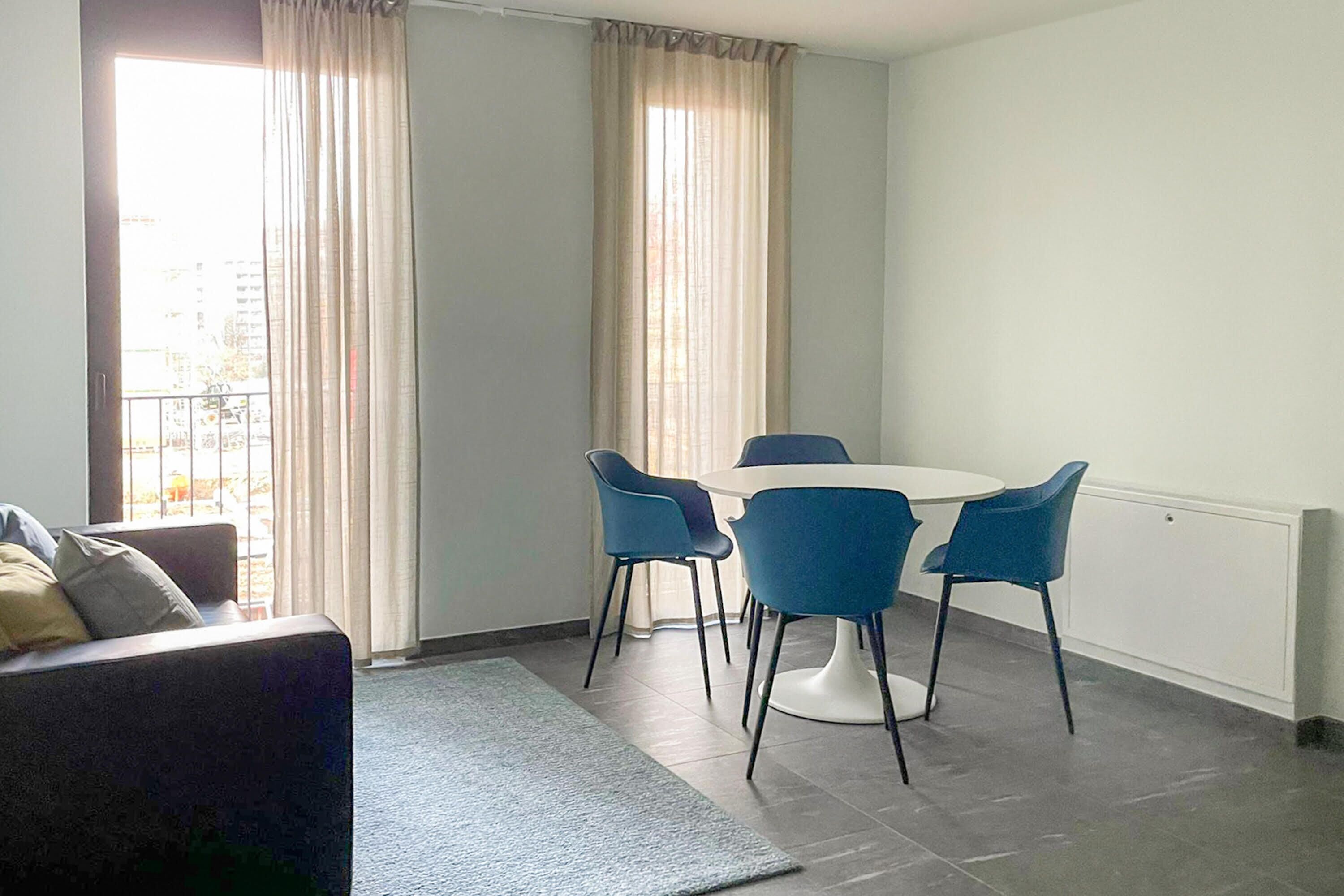 A modern living room with a sofa, a dining table with four chairs, and a large window with curtains.