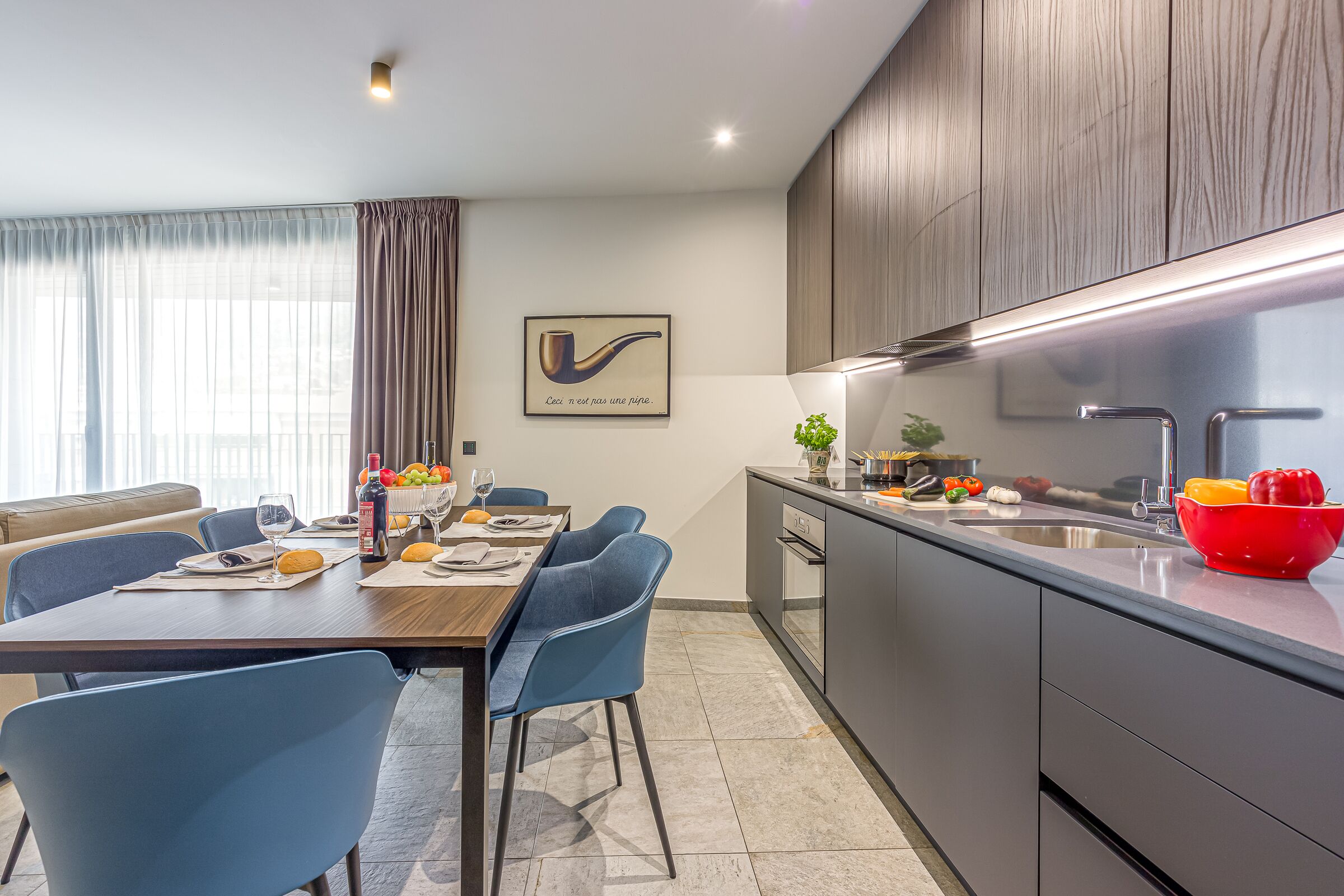 Une cuisine moderne avec un coin repas, comprenant une table en bois, des chaises bleues et un saladier rouge posé sur le comptoir.