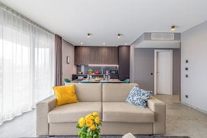 Ein modernes Wohnzimmer mit einem beigefarbenen Sofa, gelb-blauen Kissen und einer Vase mit gelben Blumen. Im Hintergrund ist eine Küche mit einem Esstisch für vier Personen zu sehen.