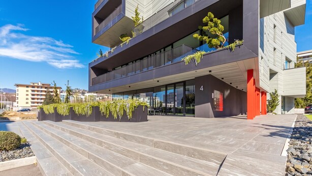 Um edifício moderno de vários andares com varandas, um caminho de pedra e uma parede de destaque vermelha.