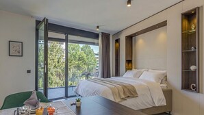 Une chambre d’hôtel moderne avec un lit, une table à manger avec des aliments pour le petit-déjeuner, une chaise verte et une vue sur les arbres à travers une grande fenêtre.