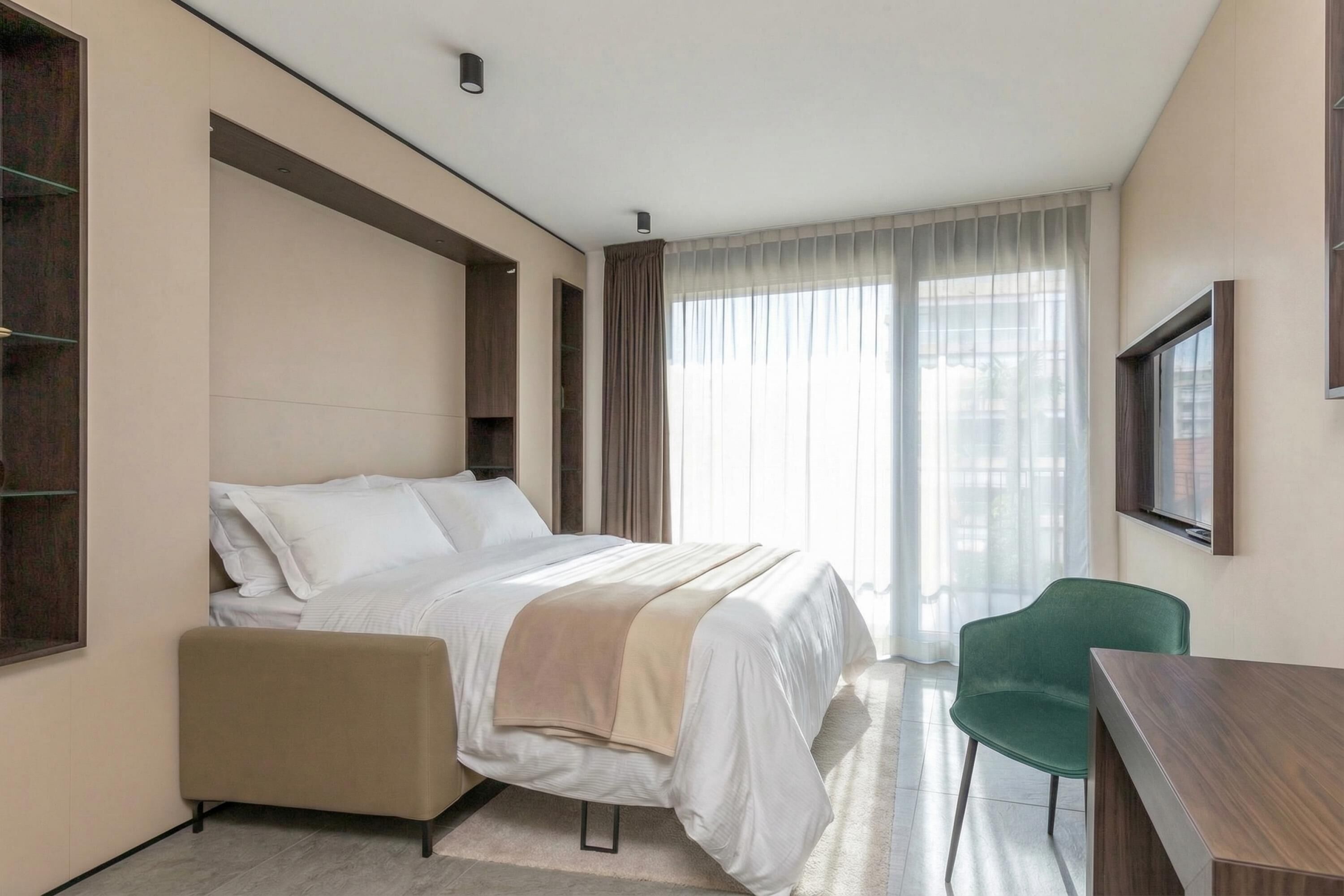 Une chambre d’hôtel moderne, équipée d’un lit, d’un fauteuil vert, d’un bureau en bois et d’une grande fenêtre avec des rideaux.