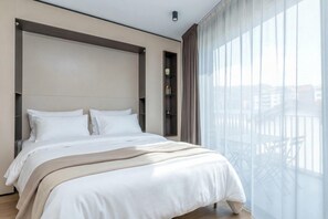 Quarto de hotel moderno com uma cama grande, prateleira embutida e vista da cidade através de cortinas leves.