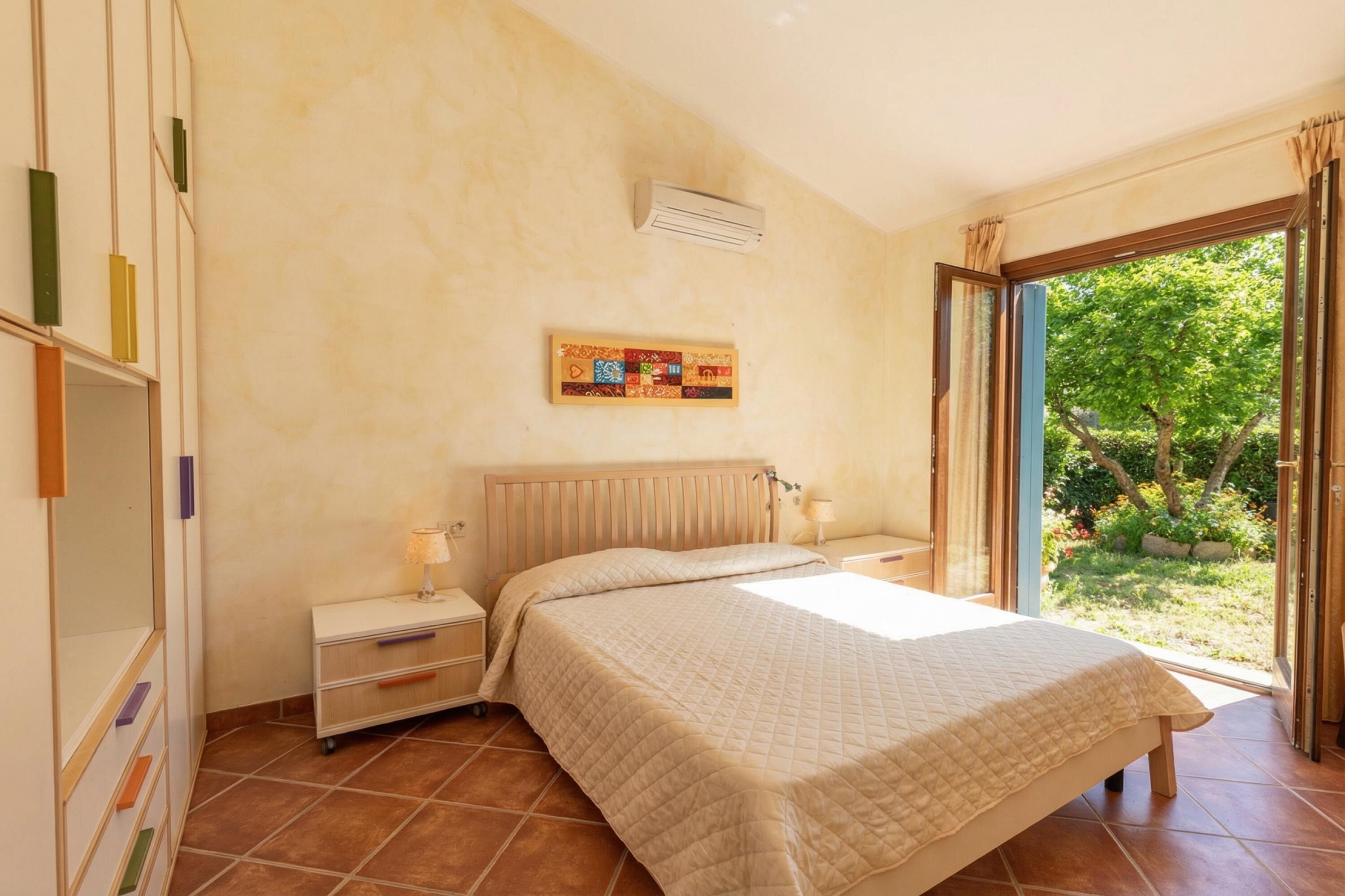Un dormitorio con cama, mesitas de noche, ventana con vistas a la vegetación y aire acondicionado de pared.
