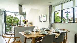 Un comedor con una mesa de madera dispuesta para cuatro personas, rodeada de sillas, una cocina al fondo y vistas a la vegetación exterior.