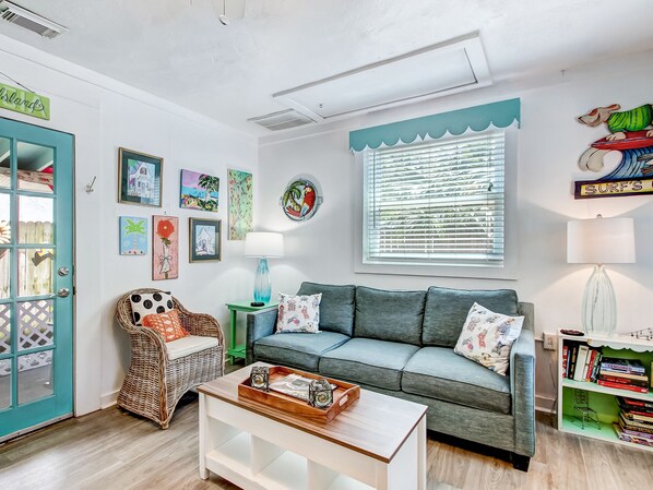 Una sala de estar con una puerta verde azulado, un sofá gris, un sillón de mimbre, una mesa de centro blanca y una estantería.