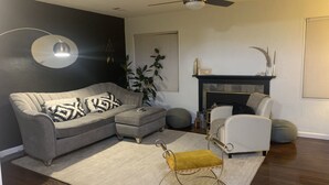 Ein Wohnzimmer mit einem grauen Ecksofa, einem weißen Sessel, einem gelben Hocker, einer Stehlampe, einem Kamin und einem Deckenventilator.