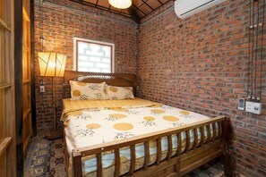 Una habitación con pared de ladrillo, cama de madera con cabecera, lámpara de pie y ventana con persianas.