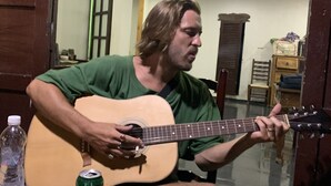 En person spiller akustisk guitar ved et bord med en flaske, en dåse og en servietæller.