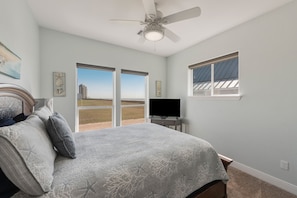 A bedroom with a bed, a ceiling fan, a TV, and a large window with a view of the outdoors. - Family Friendly Beach House in Gated Beach Resort (Galveston)