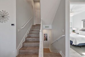 Uma escadaria com degraus de madeira e corrimão branco que leva a um quarto com uma cama e uma mesa de cabeceira.