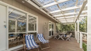 Ein überdachter Außenbereich mit Holzboden, zwei blau gestreifte Lounge-Sessel, ein Tisch mit Stühlen und große Fenster.