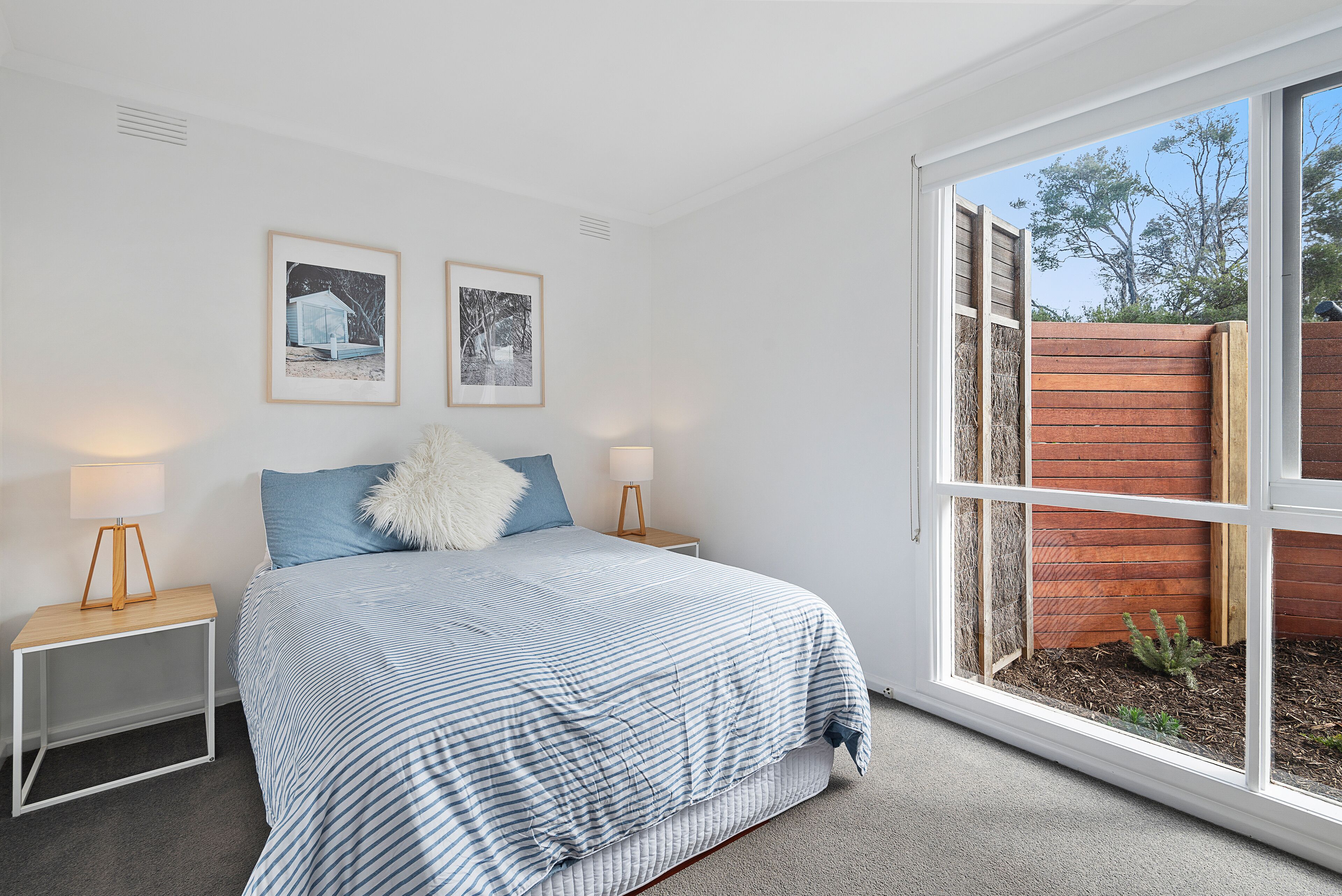A bedroom with a large window, a bed with a striped bedspread, two bedside tables with lamps, and two framed pictures on the wall.