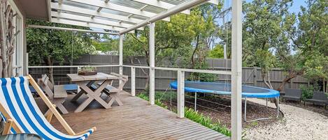 A covered outdoor deck with a table and chairs, a trampoline, and a wooden fence in the background.