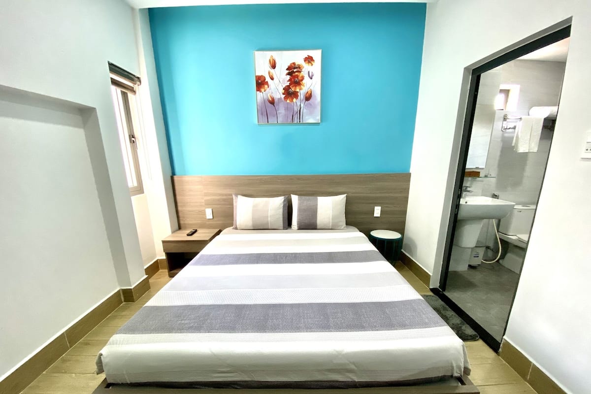 Habitación de hotel con una cama grande, una mesita de noche, una mesa redonda, un cuadro en la pared y un baño visible a través de una puerta abierta.