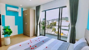 A hotel room with a large bed, a potted plant, a wardrobe, and a view of the city through a large window.
