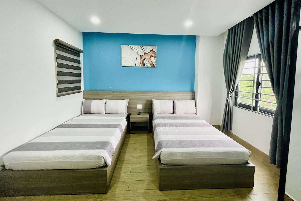 Habitación de hotel con dos camas, una pared de acento azul, un cuadro y una ventana con cortinas.