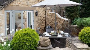 A stone building with a glass door, a patio table set with plates and a bottle, surrounded by greenery and a white umbrella. - The Nook - Guiting Power, Cheltenham sleeping 6 guests (Guiting Power)